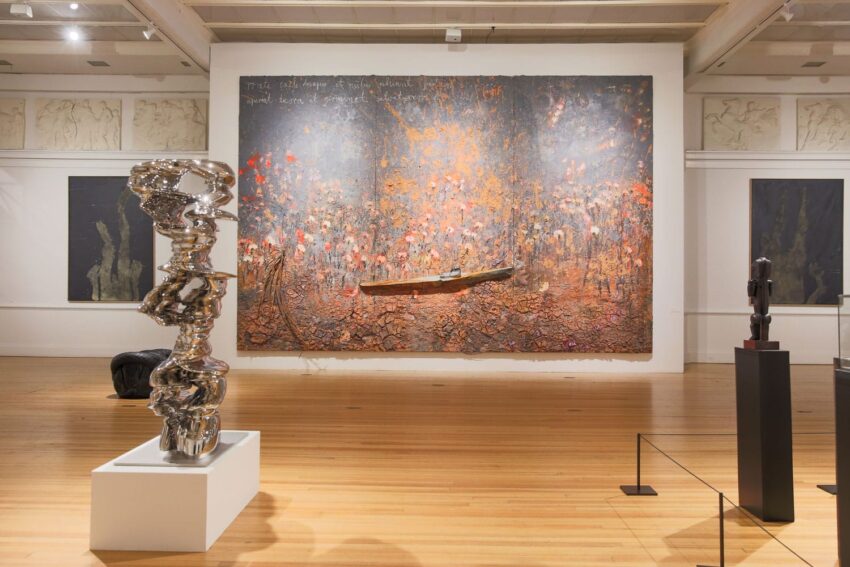 Art gallery with abstract sculpture in foreground and large textured painting on wall, featuring a boat element.