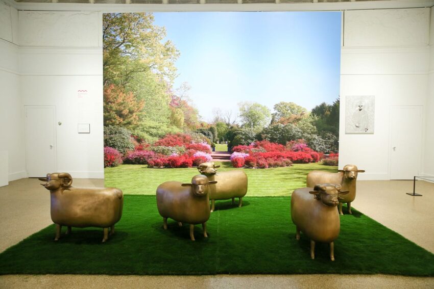 Sculpture of sheep on green grass with a backdrop of a vibrant garden scene inside a white-walled gallery.