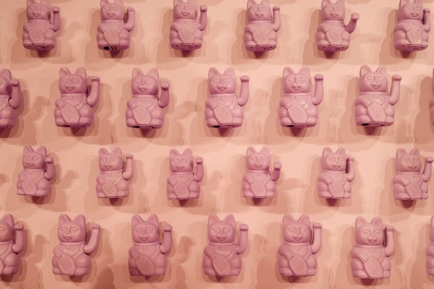 Rows of pink fortune cat statues on a wall, each with one paw raised, creating a harmonious pattern.