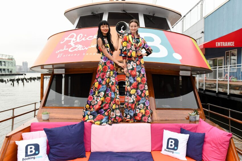 Two women in vibrant floral dresses standing on a boat deck, adorned with colorful cushions and Booking.com logos.