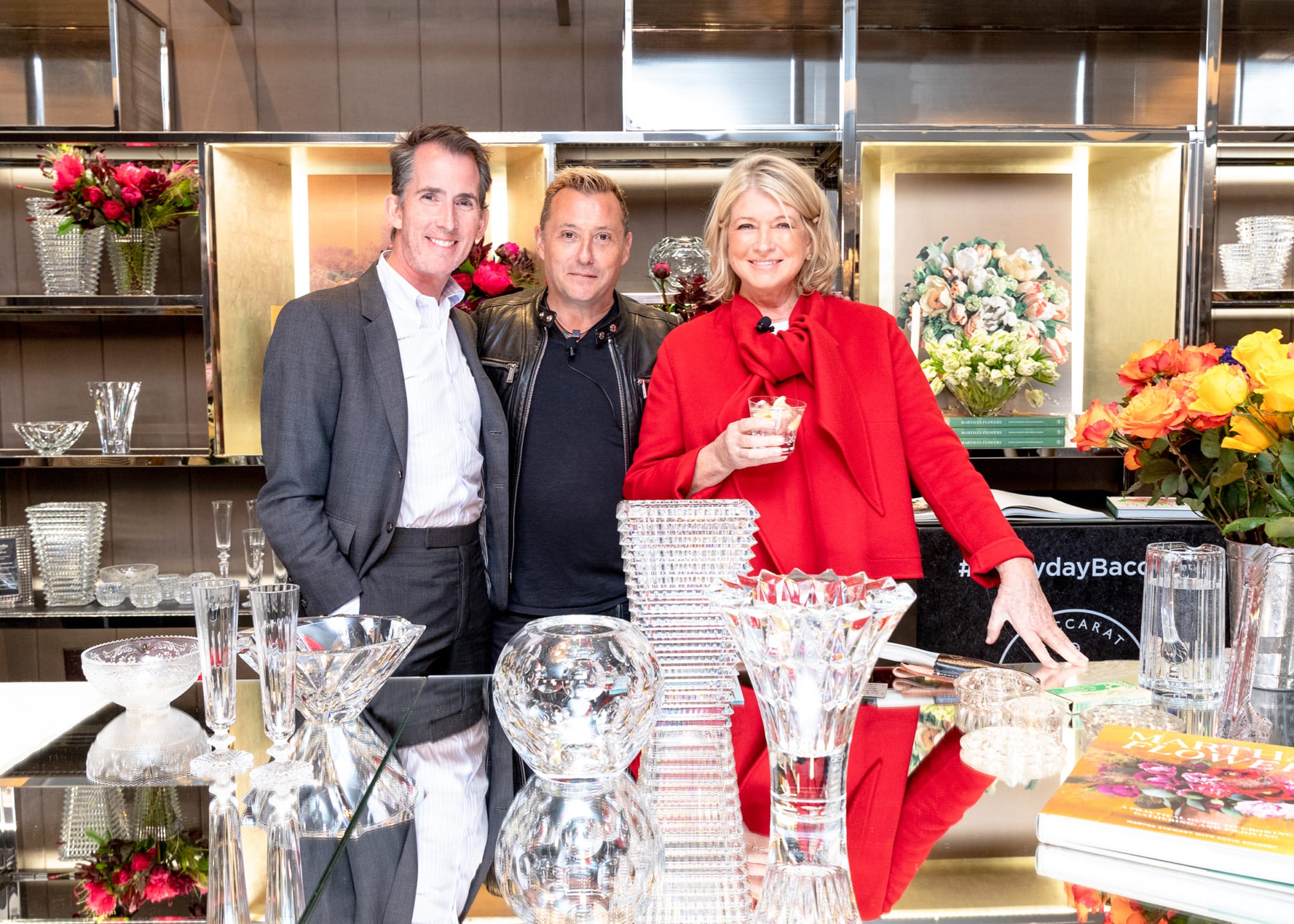 Three people standing behind a glassware display, holding drinks and smiling, with floral arrangements in the background.