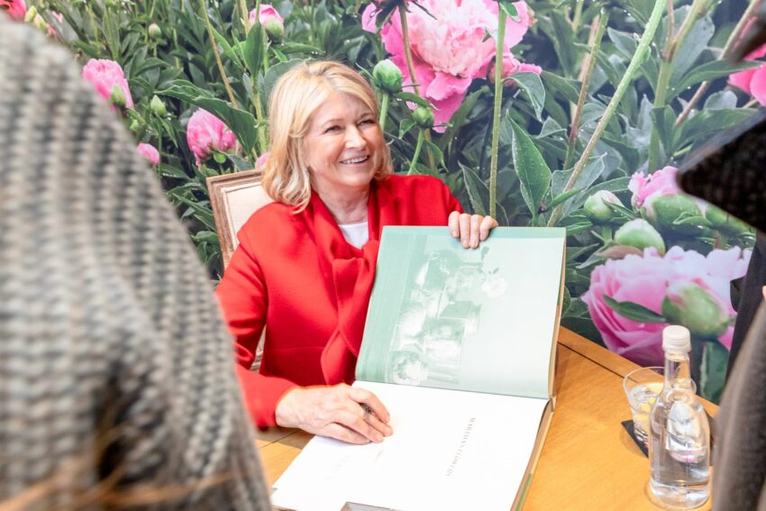 Woman in red jacket signing a book at a table, surrounded by a floral background and people nearby.