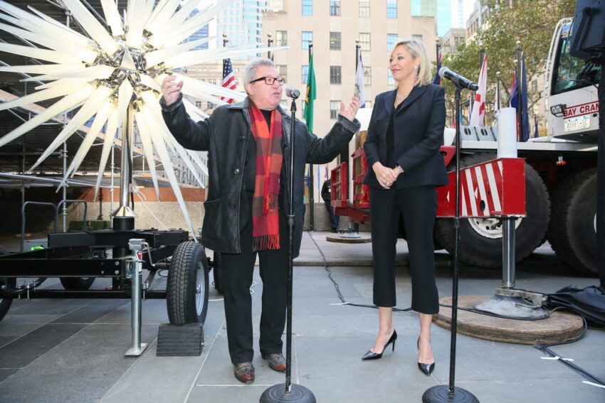 Two people speaking at a microphone near a large decorative star and a truck in an urban setting.