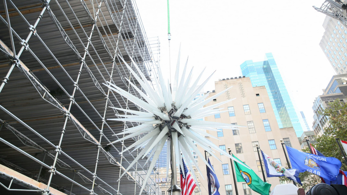 Large glass star sculpture being installed near a building with scaffolding and flags, viewed from below against a cloudy sky.