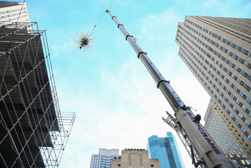 Crane raising a decorative spiky structure against a blue sky amid tall city buildings and scaffolding.