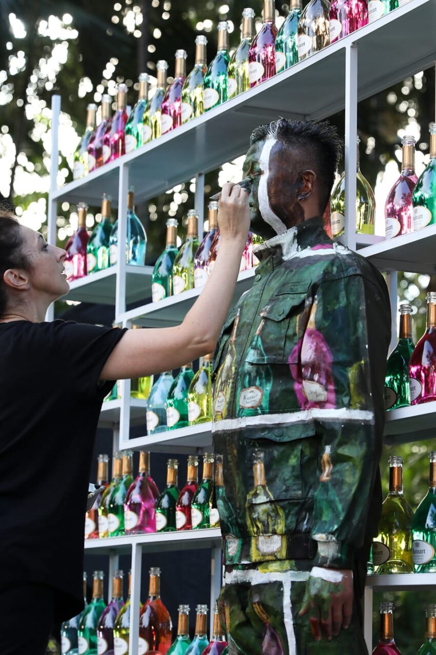 Artist painting person to blend in with colorful bottles on shelves in an outdoor setting.