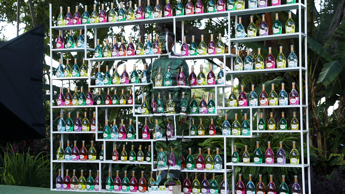 Person camouflaged among shelves filled with colorful bottles in a lush outdoor setting.