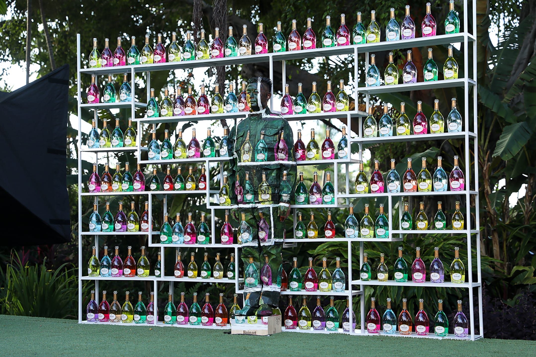 Person camouflaged among shelves filled with colorful bottles in a lush outdoor setting.