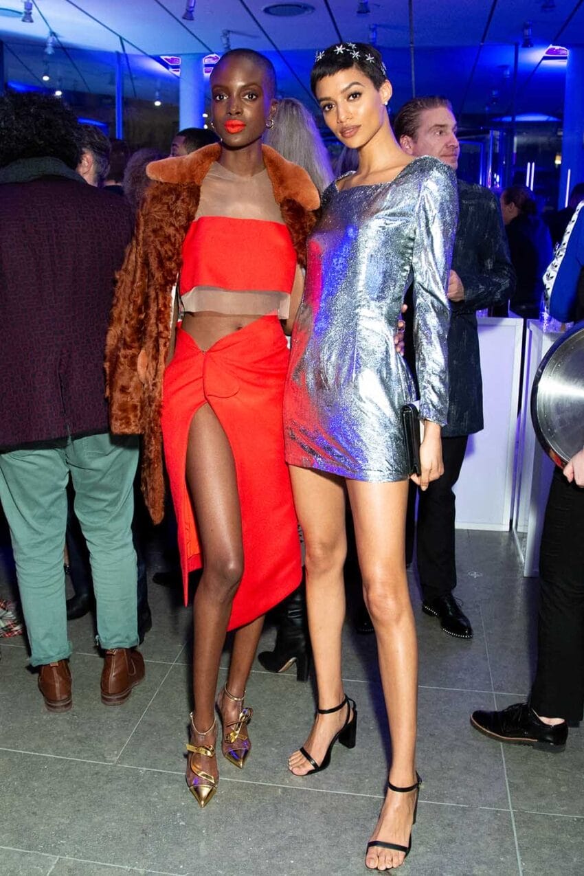 Two models in vibrant red and silver evening dresses pose at a lively event with multiple guests in the background.