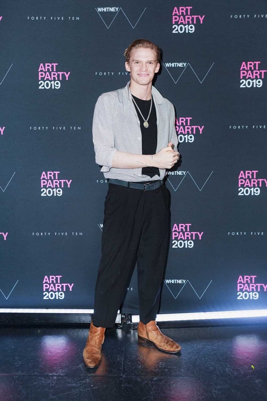 Person posing at an art party event, wearing a gray shirt and black pants with a pink and black event backdrop.