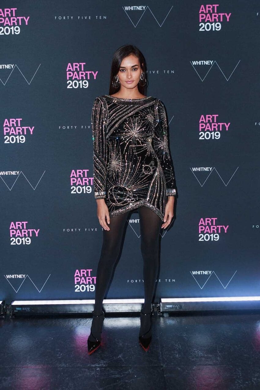 A person in a shimmering black dress poses at an art party event with a backdrop featuring the year 2019.
