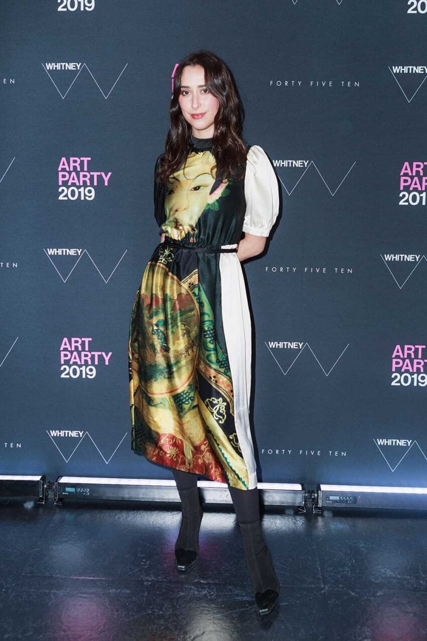 Person posing at art party event wearing a colorful patterned dress against a branded backdrop.