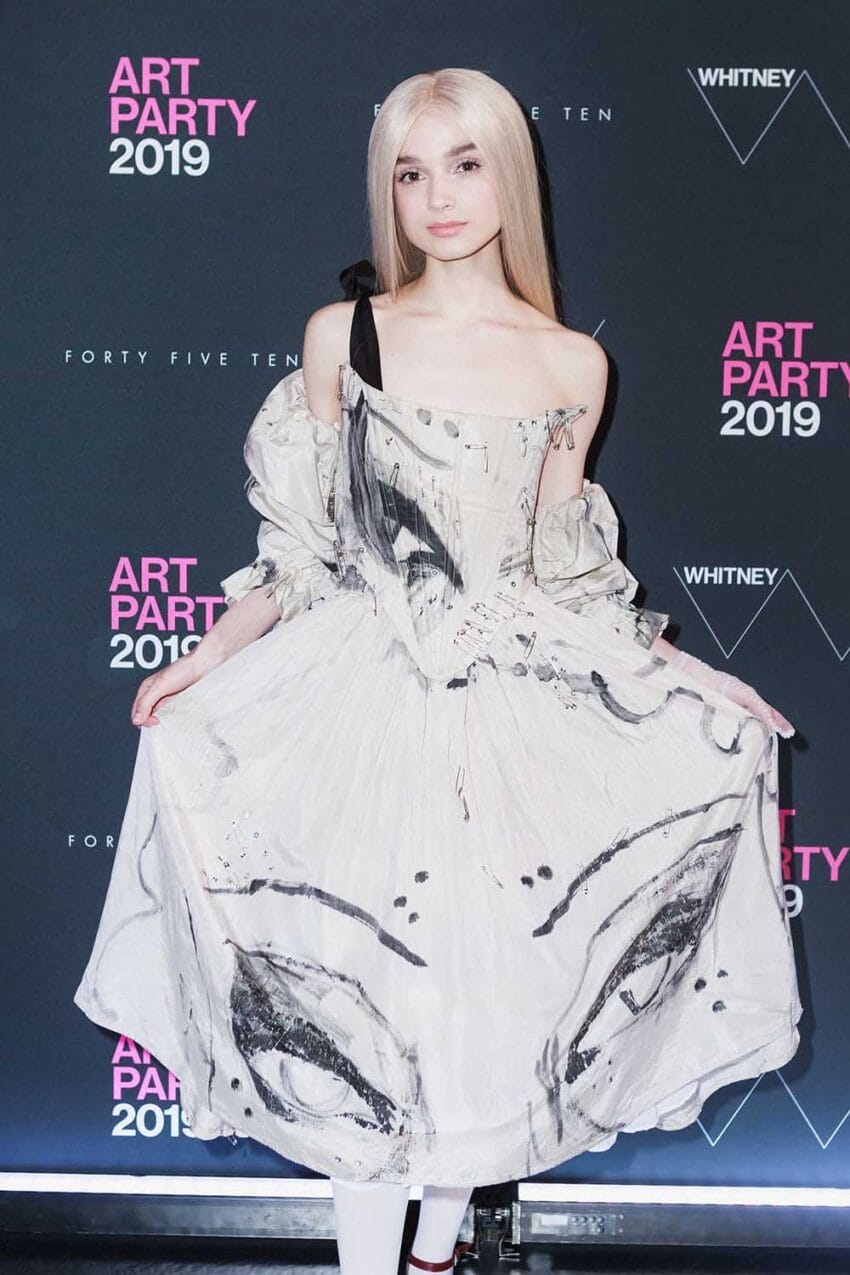 Person in a white dress with black abstract design at an art event, holding the sides of the dress, in front of a backdrop.