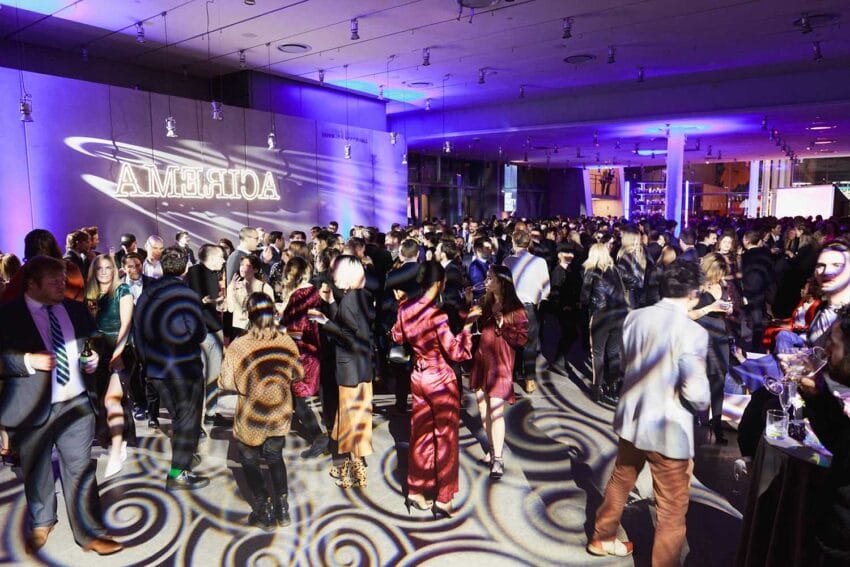 A large crowd of people at a vibrant indoor party with colorful lighting and "ACRERMA" projected on the wall.