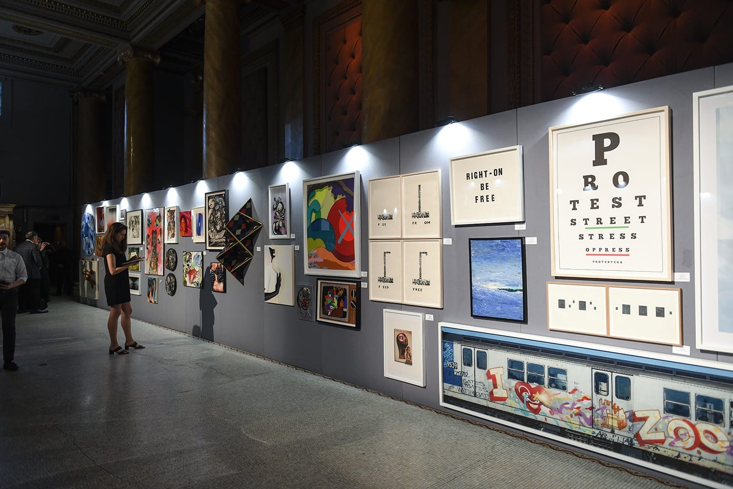 Art gallery exhibition with various framed paintings and photographs displayed on a gray wall, people observing the artwork.