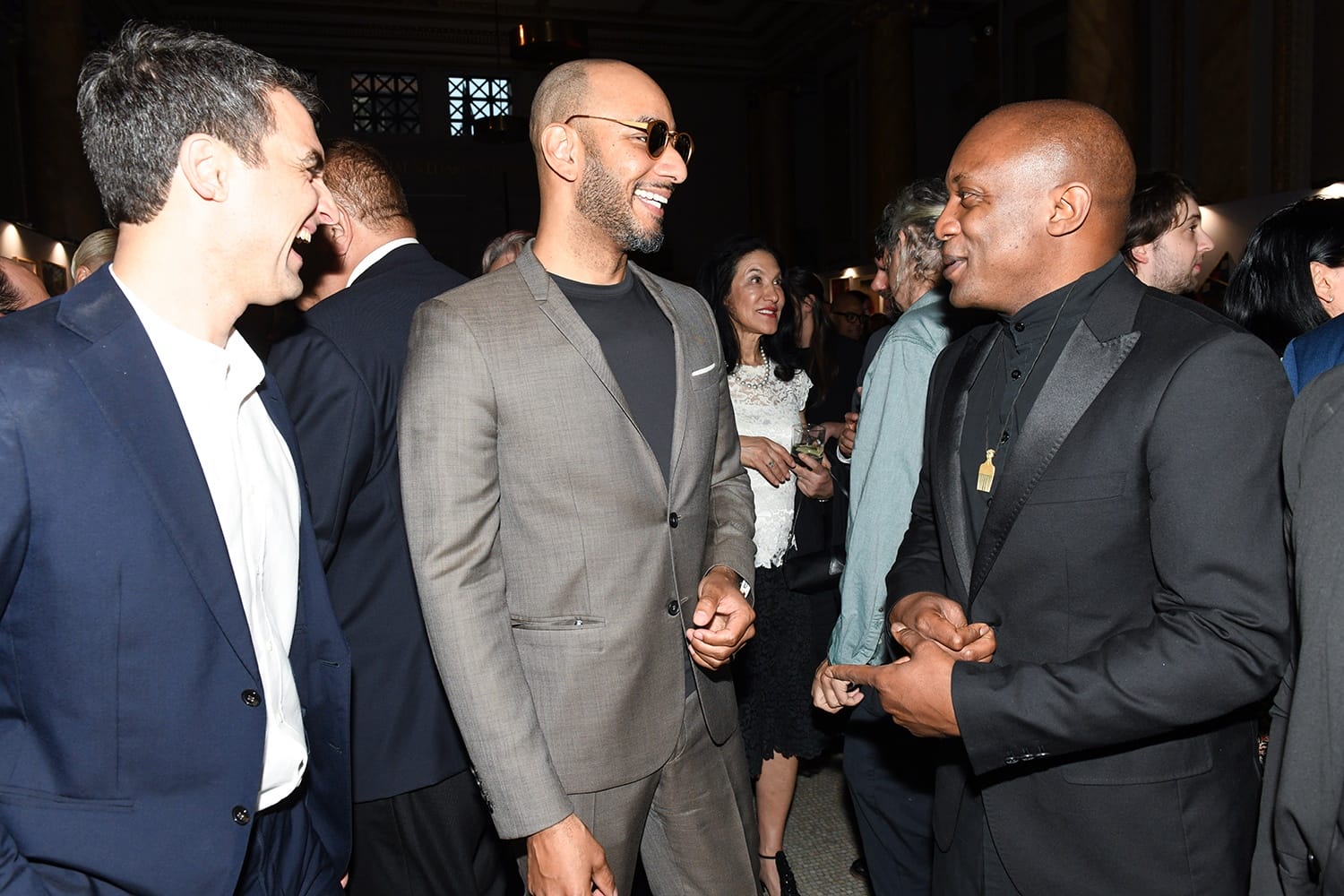 Three men in suits laughing and conversing at a formal gathering with people socializing in the background.