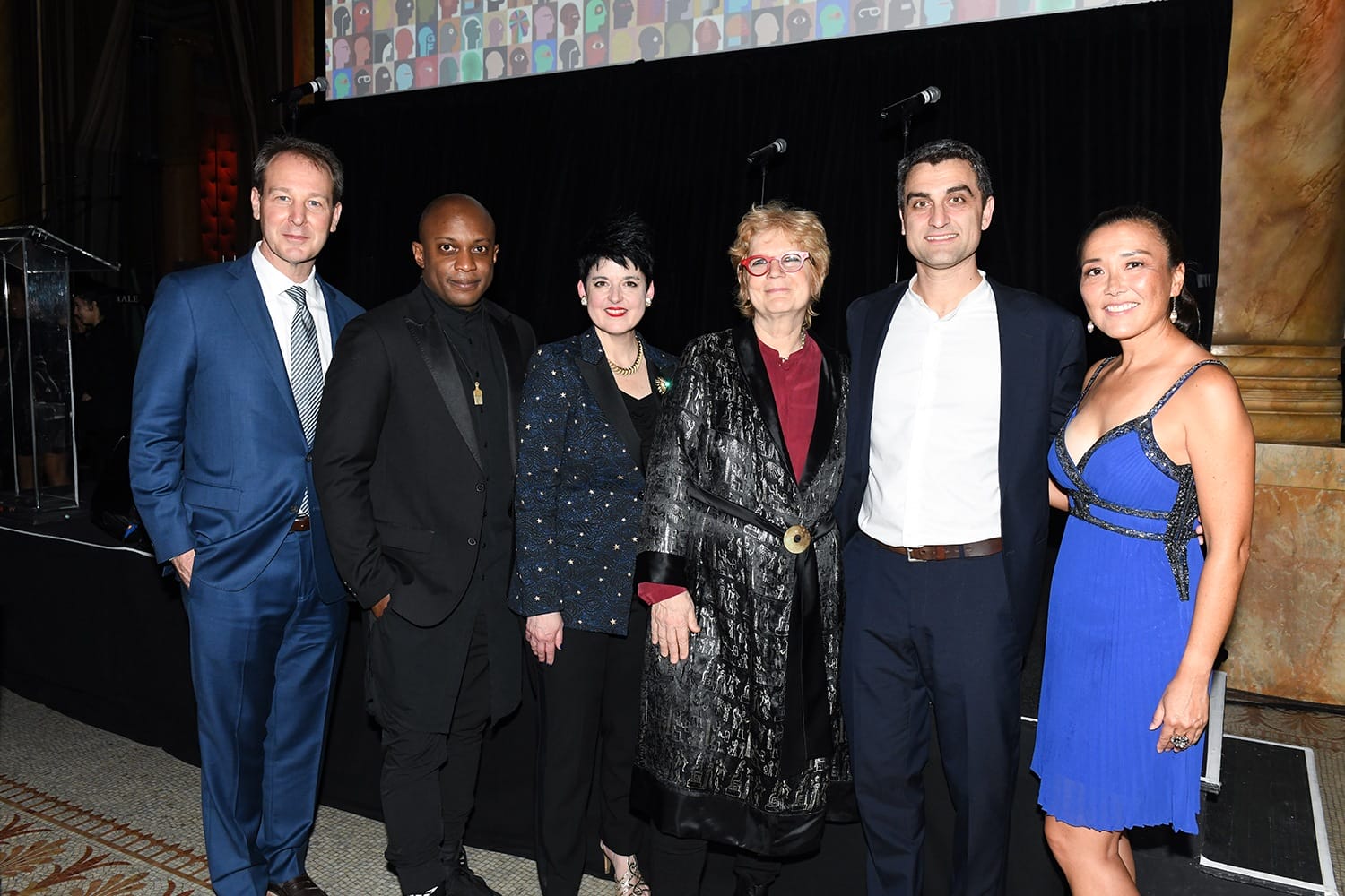 Group of six people posing for a photo on stage, dressed formally, against a backdrop with a colorful design.