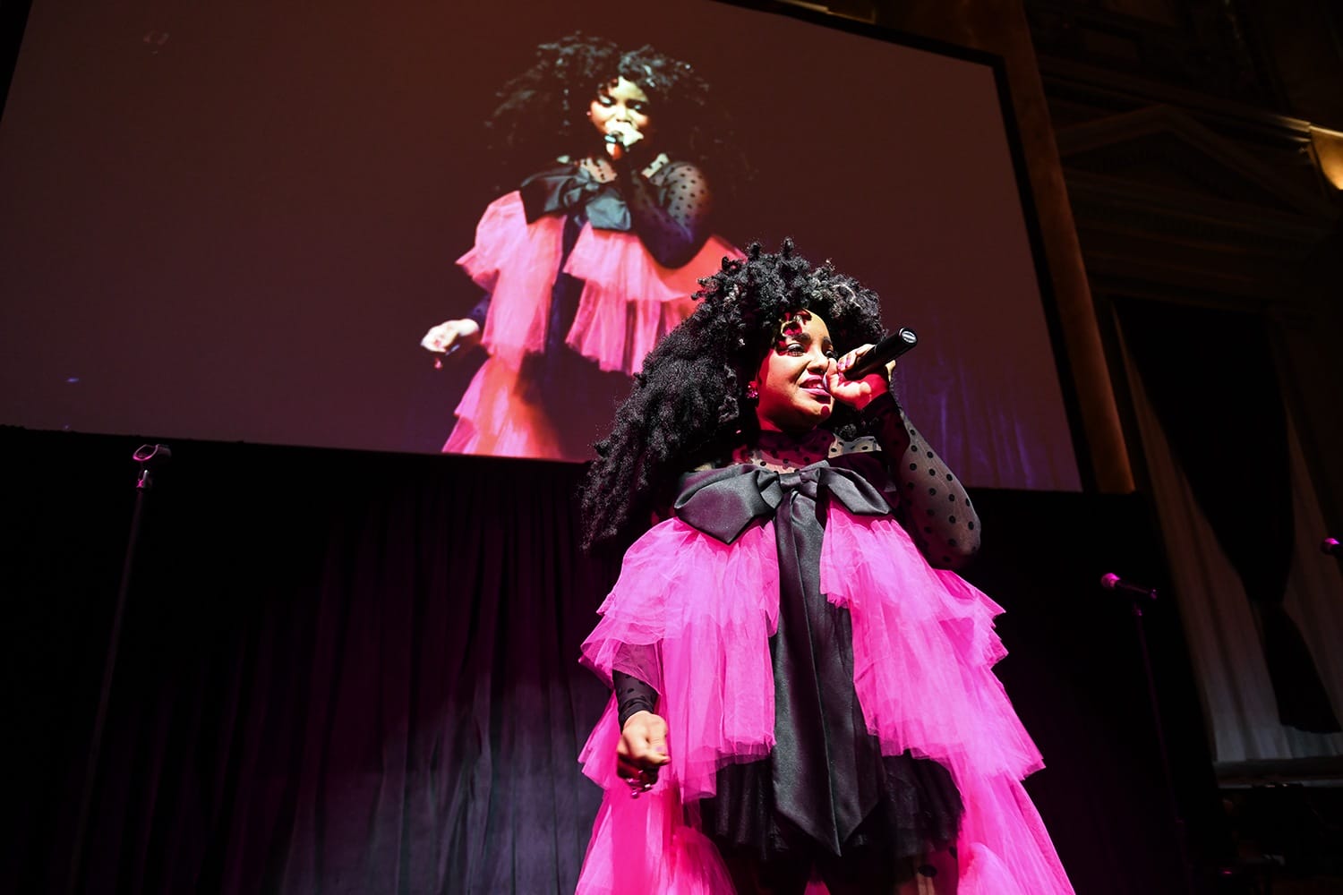 Performer in a vibrant pink dress singing on stage with their image projected on a large screen behind them.