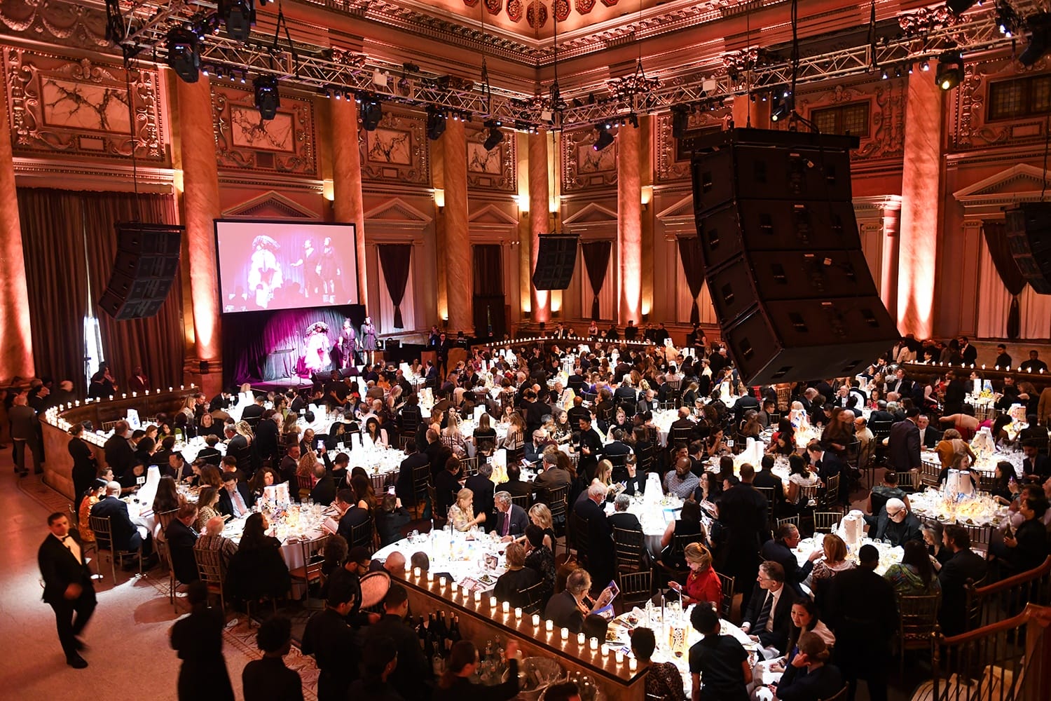 Elegant ballroom event with a large crowd seated at tables, stage with a screen, and ornate architectural details.