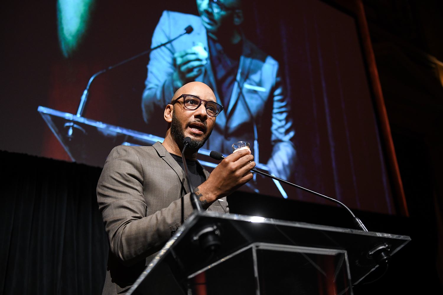 Man speaking at a podium with a large screen in the background displaying his image.