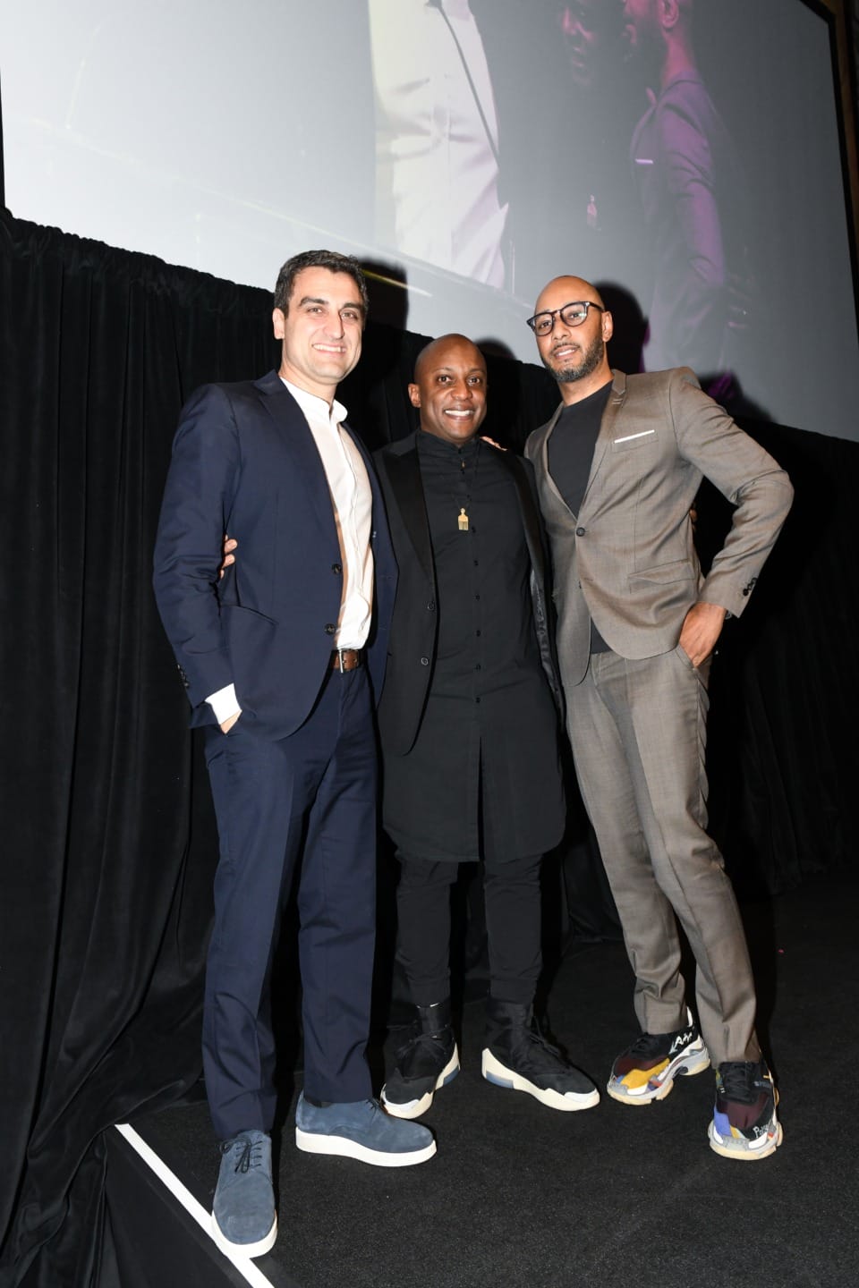 Three men in suits stand together posing for a photo on stage with a black curtain backdrop.