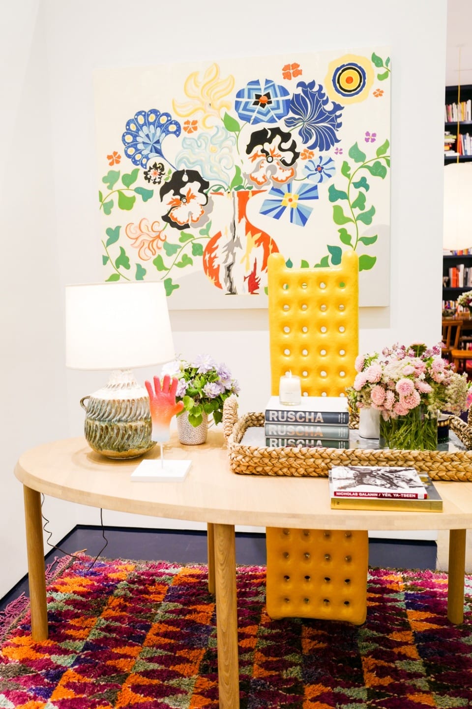 Colorful office with yellow chair, wooden desk, vibrant rug, books, flowers, abstract wall art, and modern table lamp.