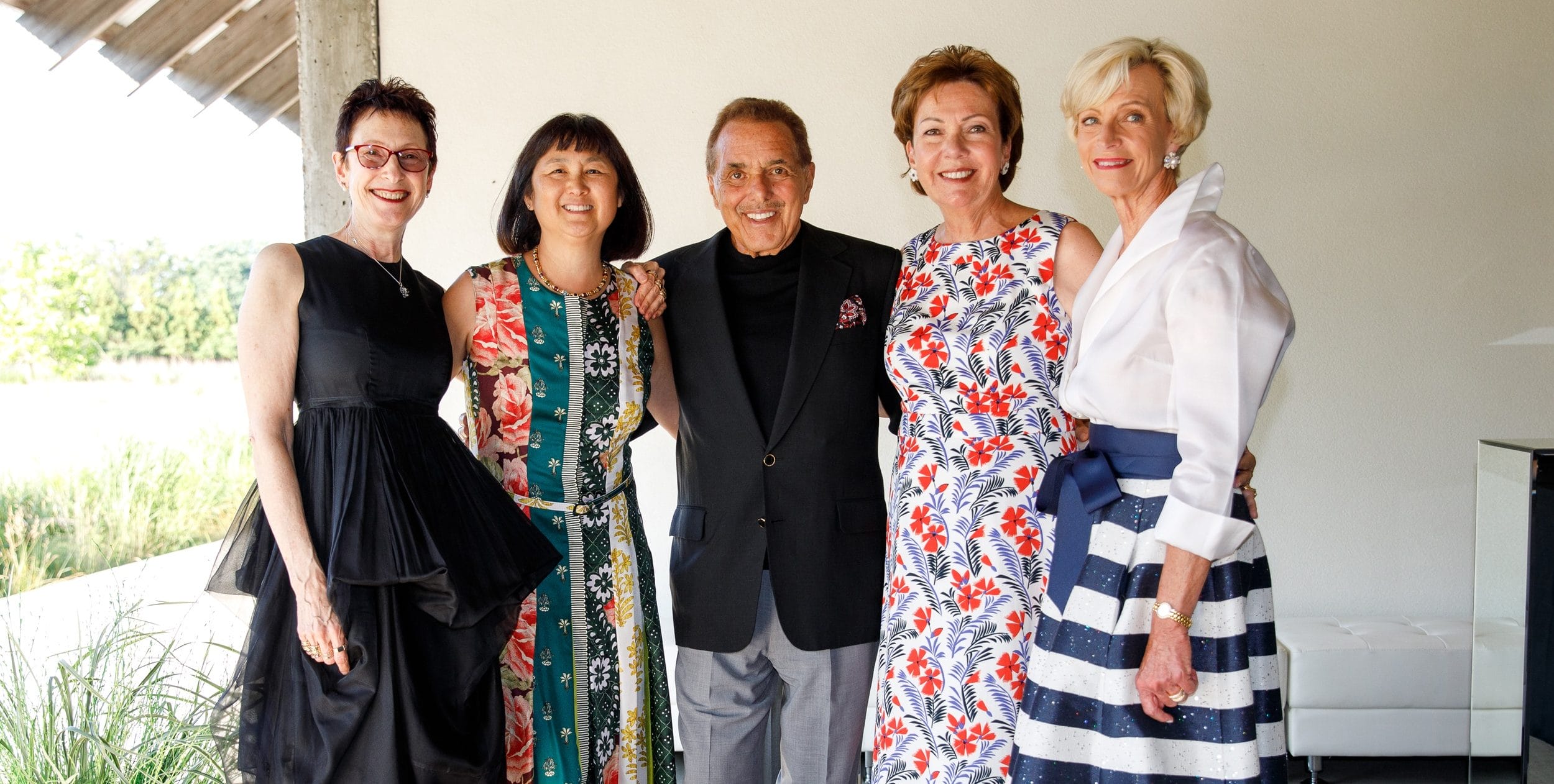 A group of five people standing together, smiling warmly at an outdoor gathering.