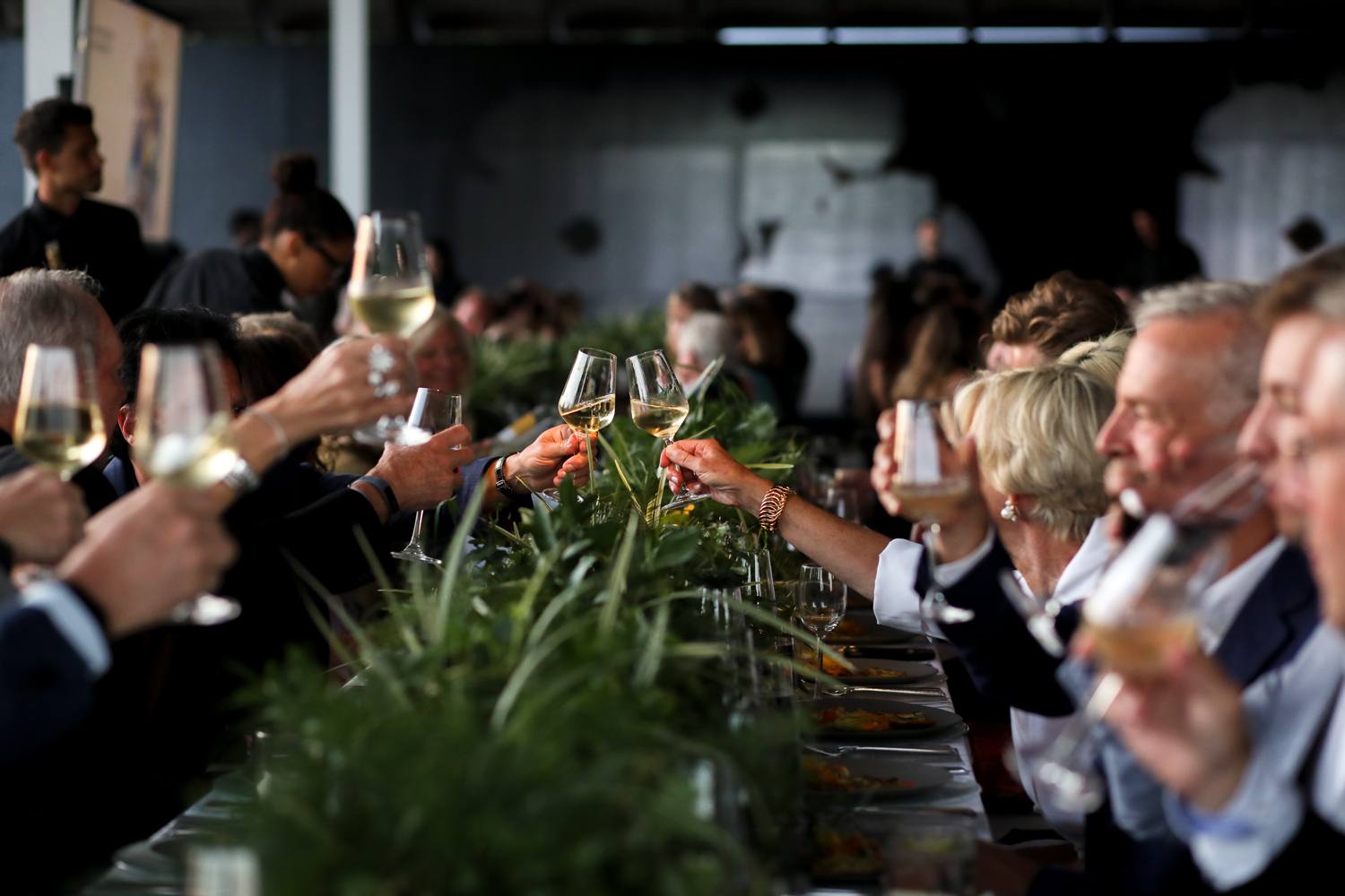 People toasting with wine glasses at a long table decorated with greenery, celebrating at a social gathering.
