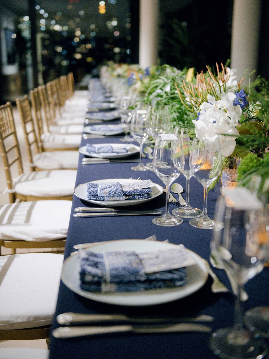 Elegant table setting with blue tablecloth, arranged plates, napkins, glasses, and a floral centerpiece in a dimly lit room.