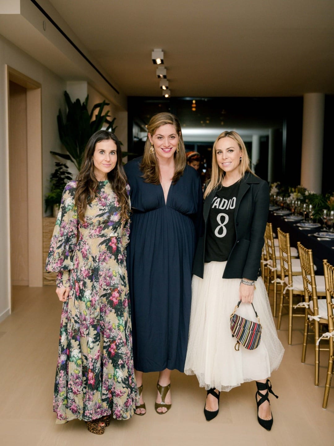 Three women posing indoors, two in floral dresses and one in a black top and white skirt, with gold chairs in the background.