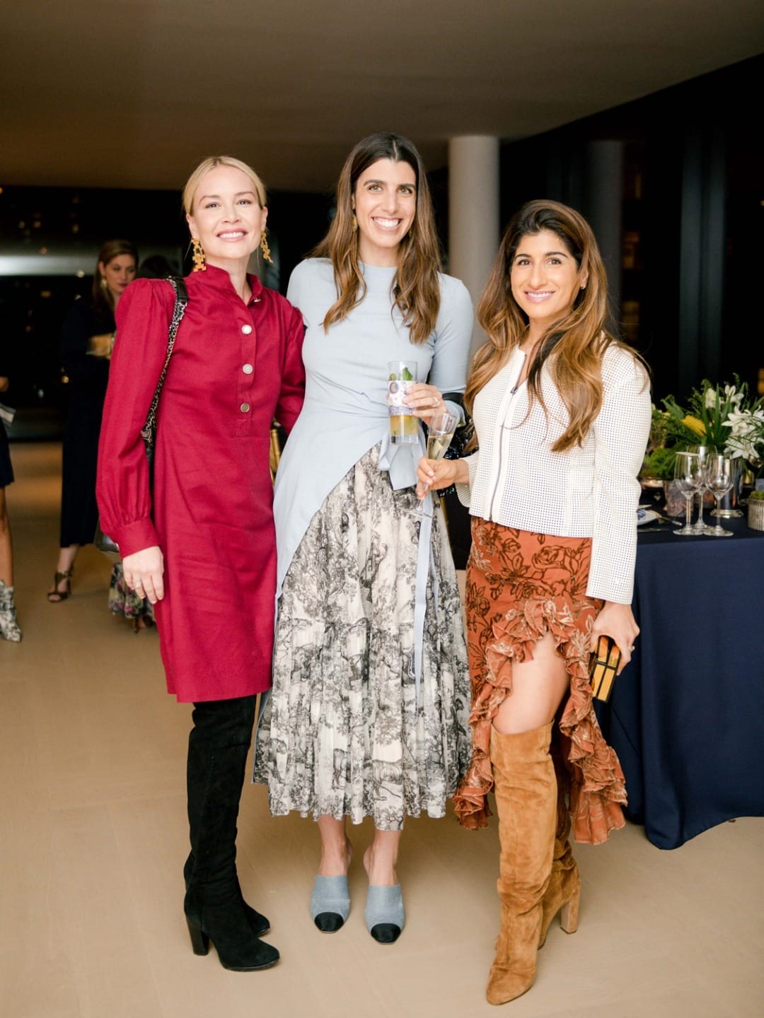 Three women smiling at a social event, wearing stylish outfits, standing in a well-lit room with tables and drinks in the background.