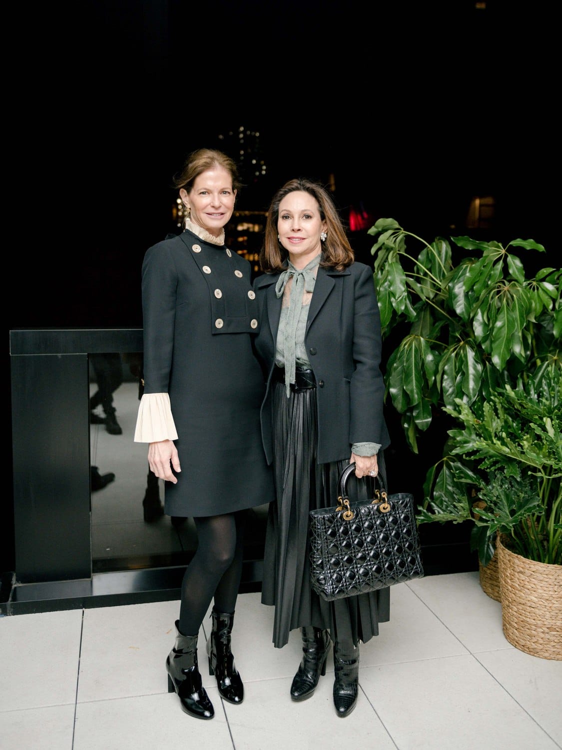 Two women in elegant black outfits standing together indoors, beside a large plant.