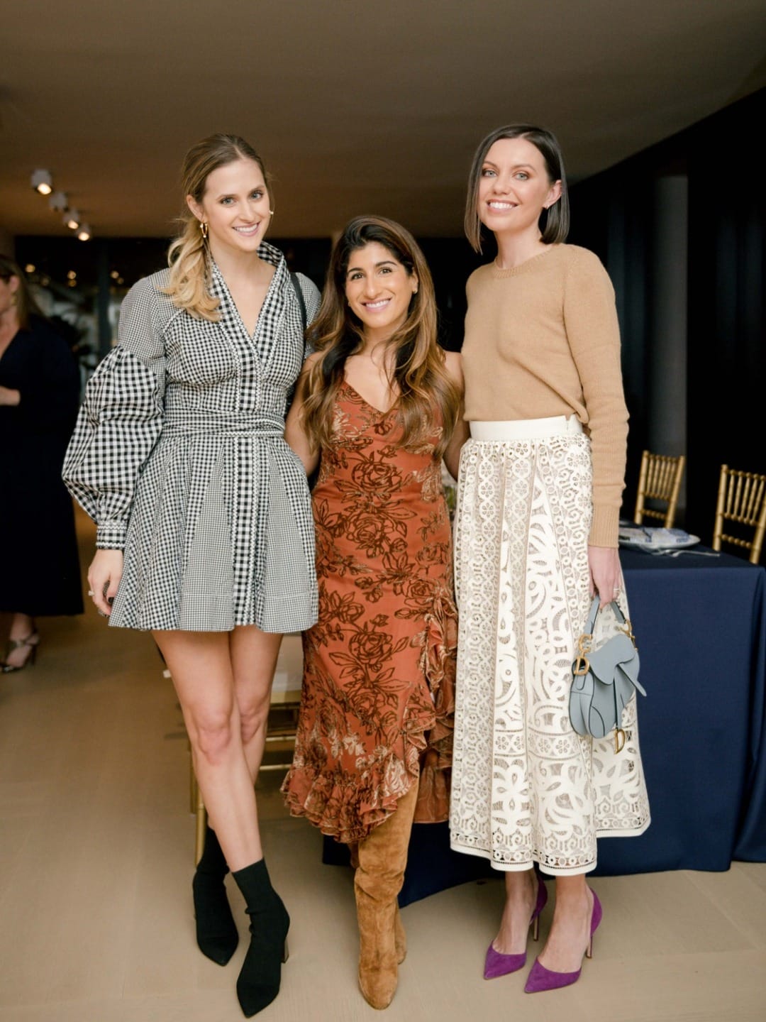 Three women smiling and posing together at an indoor event, dressed in fashionable outfits.