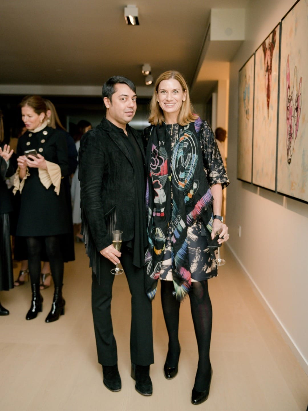 Two people standing together at an art gallery event holding champagne glasses, with colorful artwork in the background.