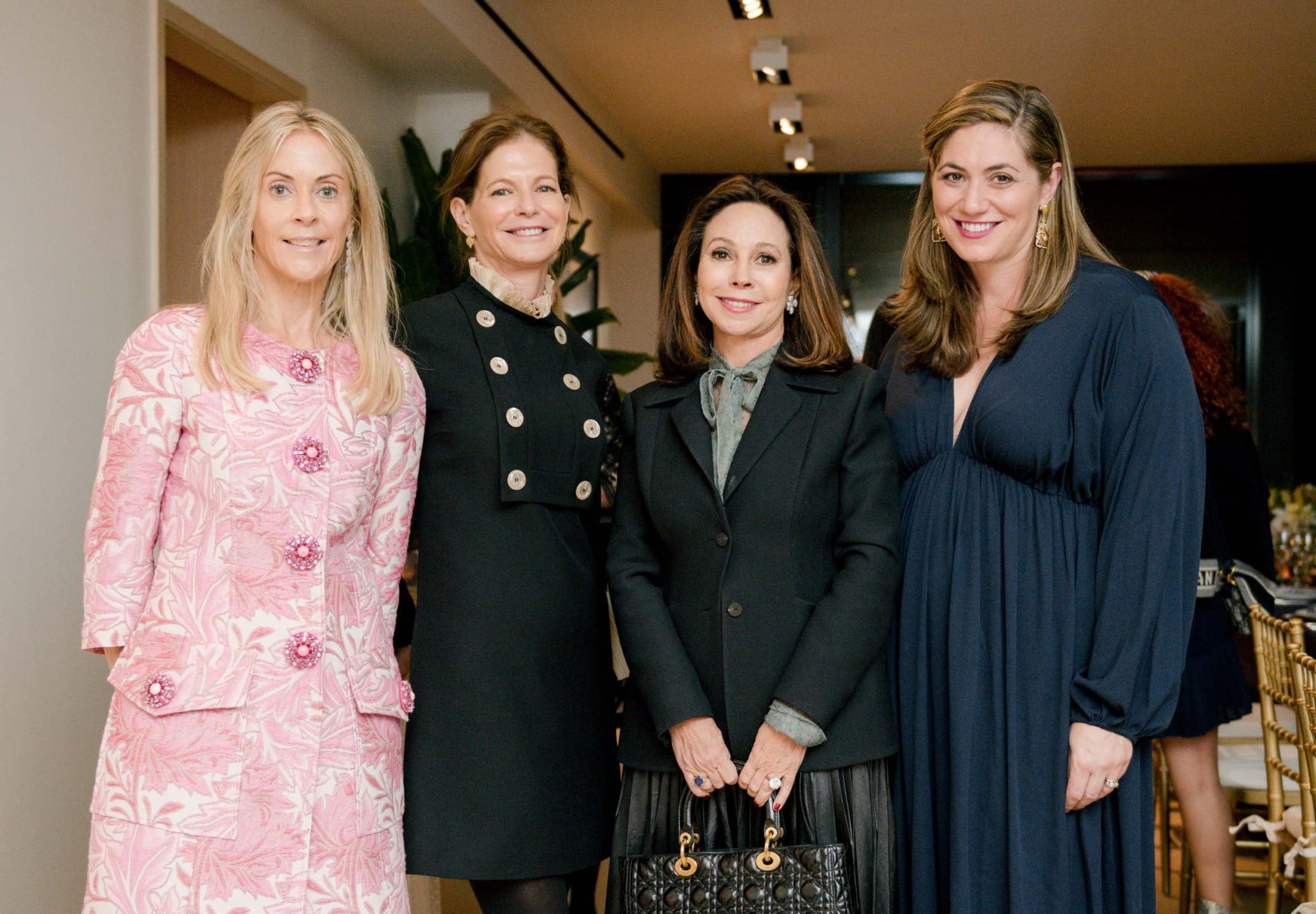 Four women standing together, smiling in formal attire at an indoor event.