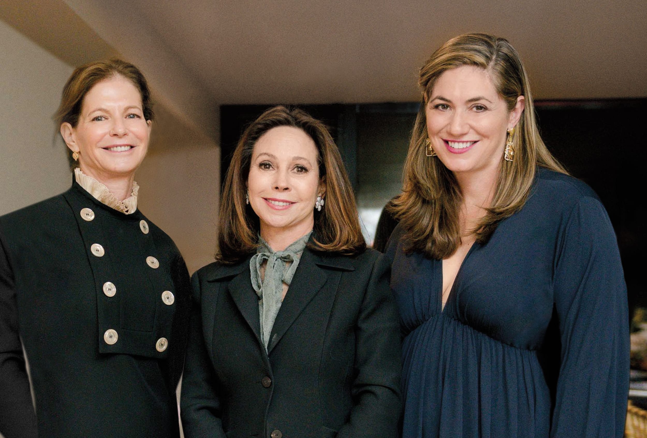 Three women smiling and posing together in formal attire indoors.