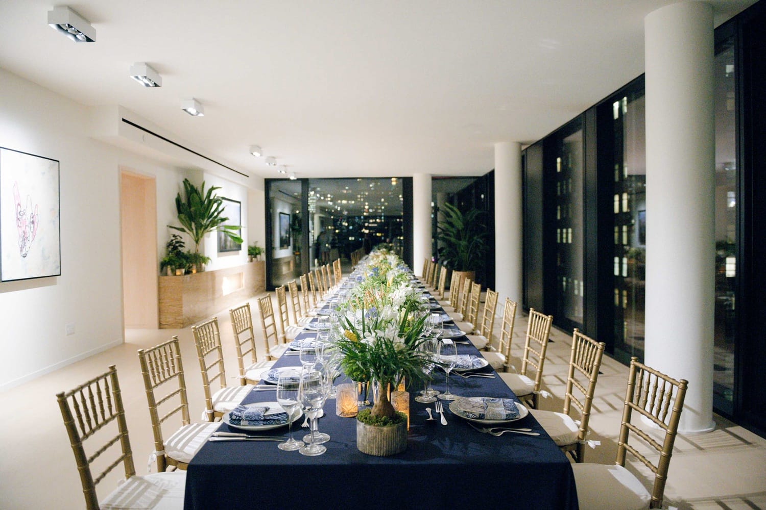 Elegant dining room with a long table set for a formal dinner, featuring gold chairs and decorative plants.