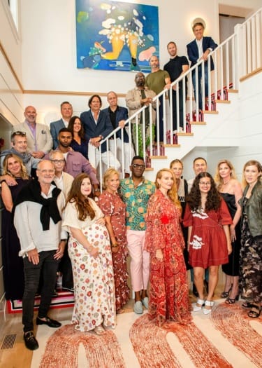 Group of people gathered on and around a staircase in a brightly lit room, with a colorful painting on the wall behind them.