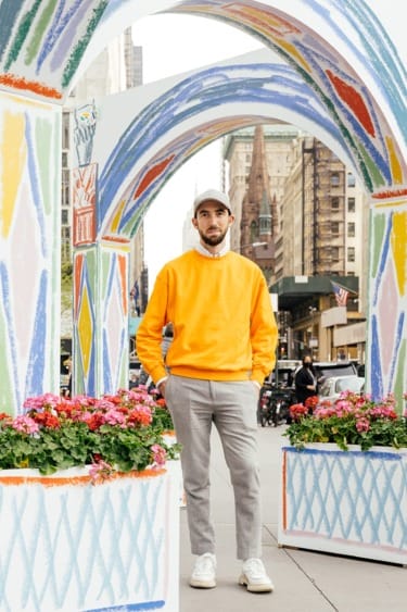 Person in a yellow sweater and gray pants standing under colorful arches on a city street with flowers and buildings.