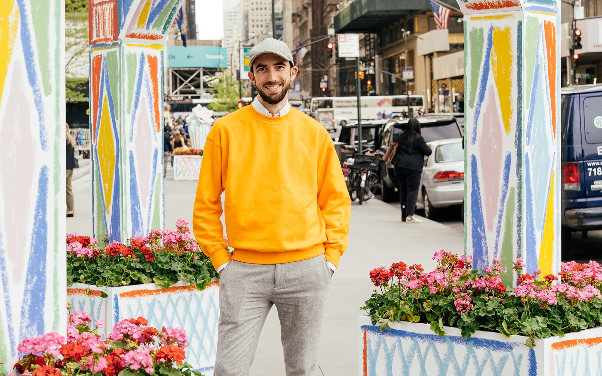Man in orange sweater standing on a colorful street with flowers and urban background.