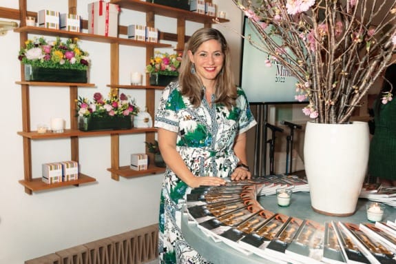 Woman in a floral dress stands near a round table with magazines, decorated with flowers and candles on shelves.