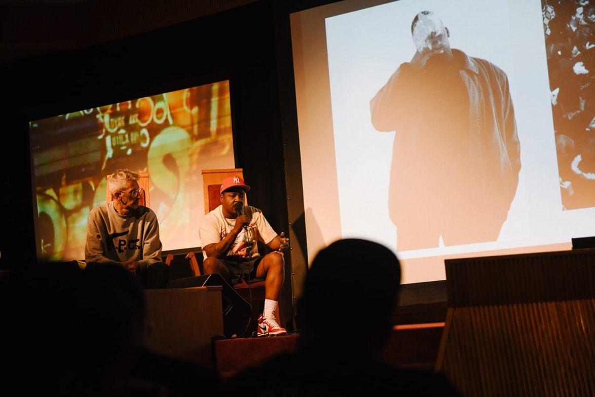 Two people sit on stage talking, with a large screen displaying black and white art beside them.