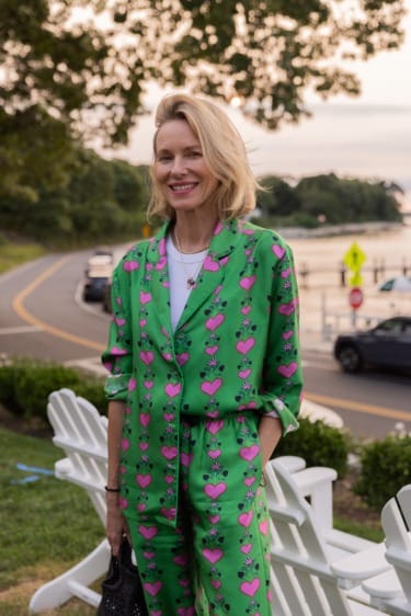 Person in a green suit with pink hearts stands outdoors near a road and benches, smiling with trees and a lake in the background.