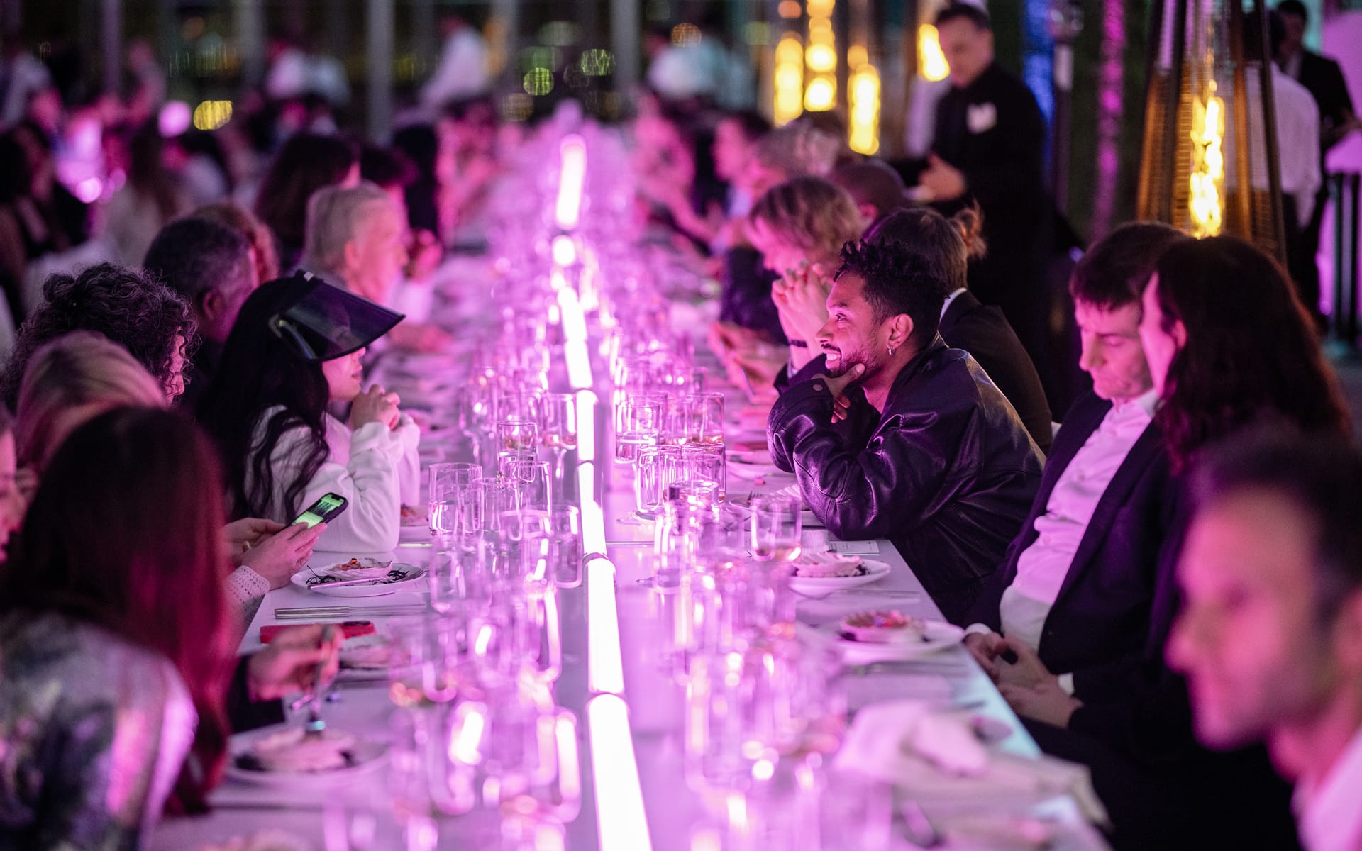 Guests sit at a long, elaborately set table with vibrant pink lighting during a festive gathering.