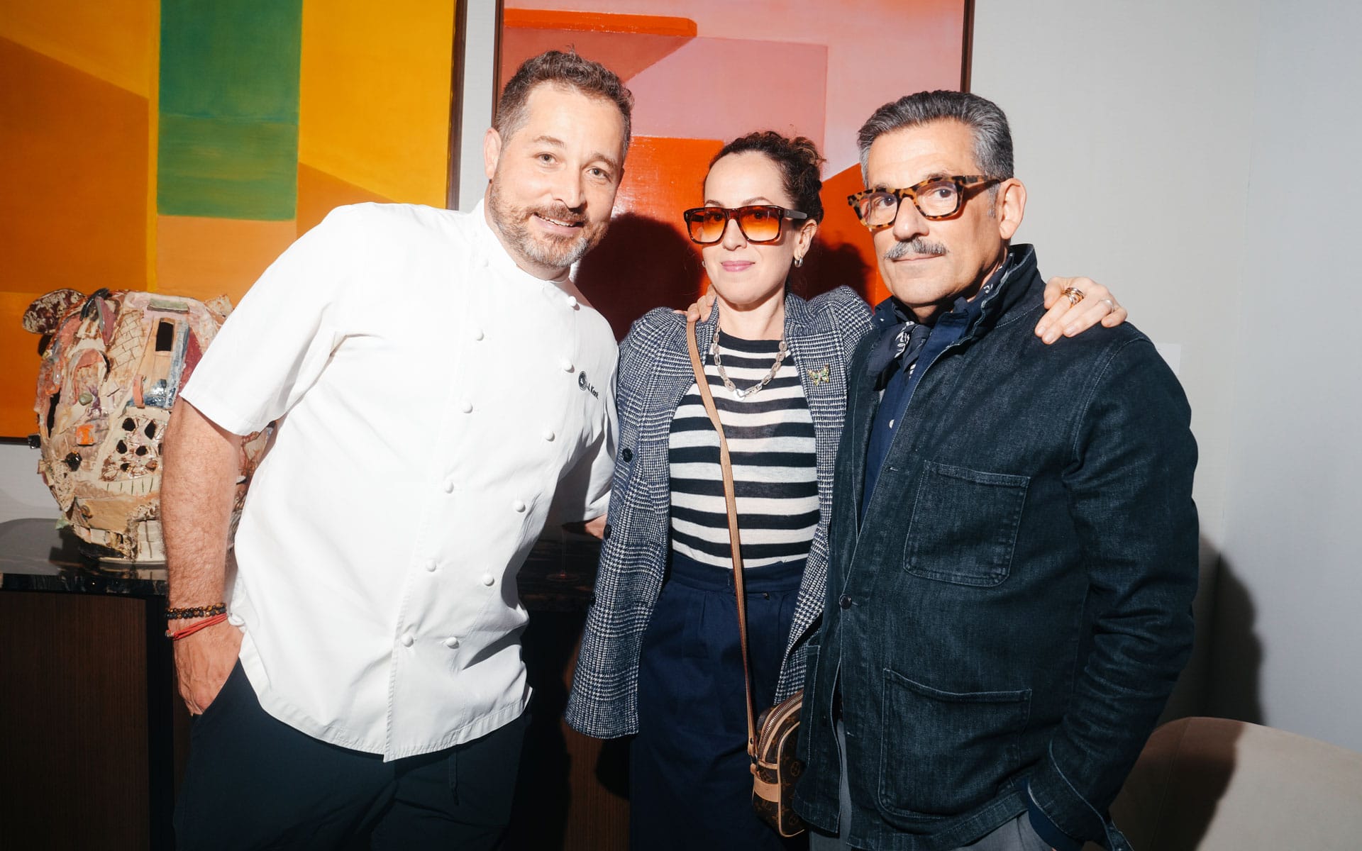 Three people standing together, smiling indoors, with colorful artwork in the background.