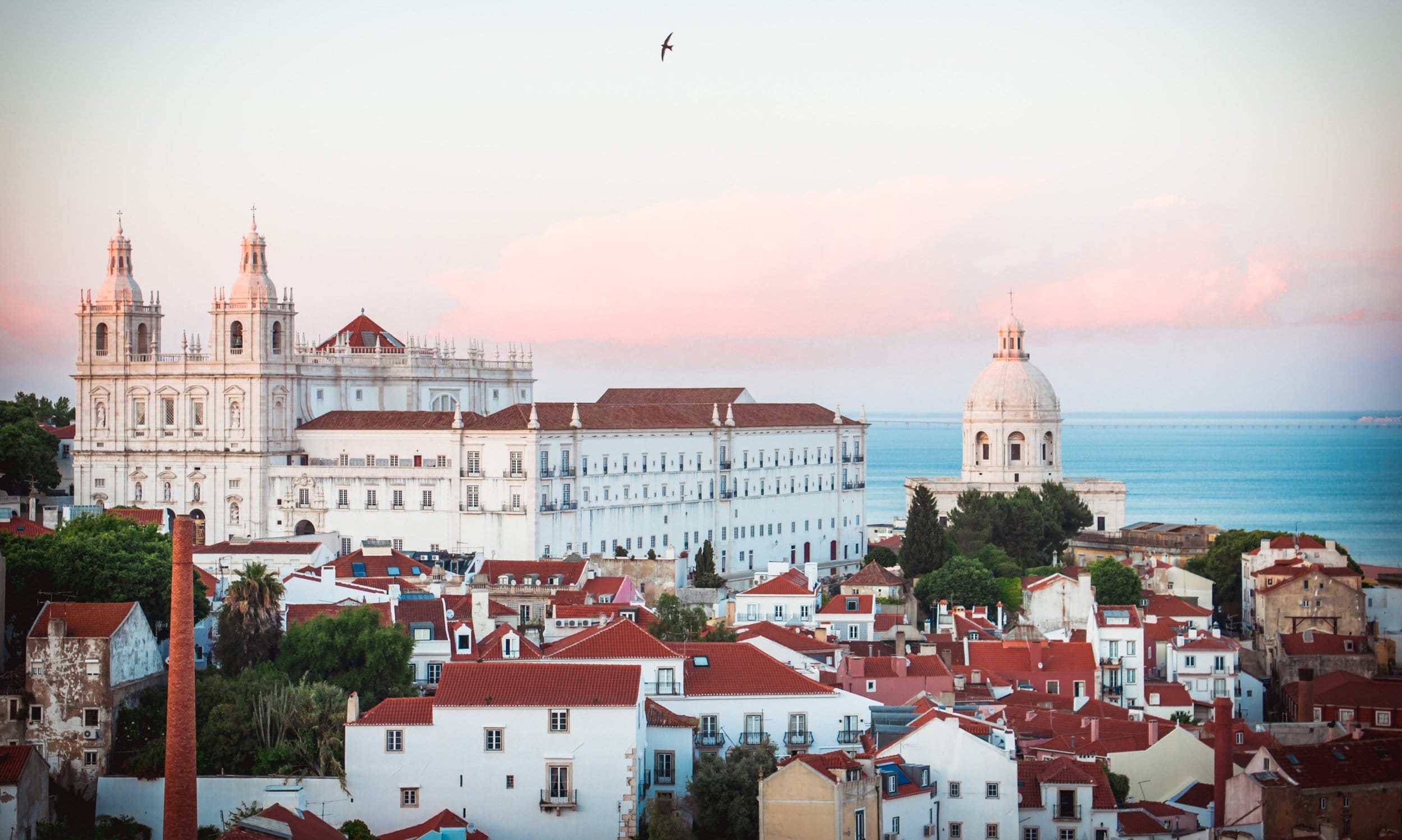 Lisbon cityscape with white monastery, red rooftops, and ocean in the background at sunset, bird flying in the sky