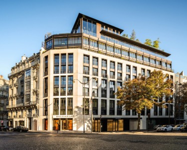 Street view of a modern, multi-story building with large windows and corner balconies, set against a clear blue sky.