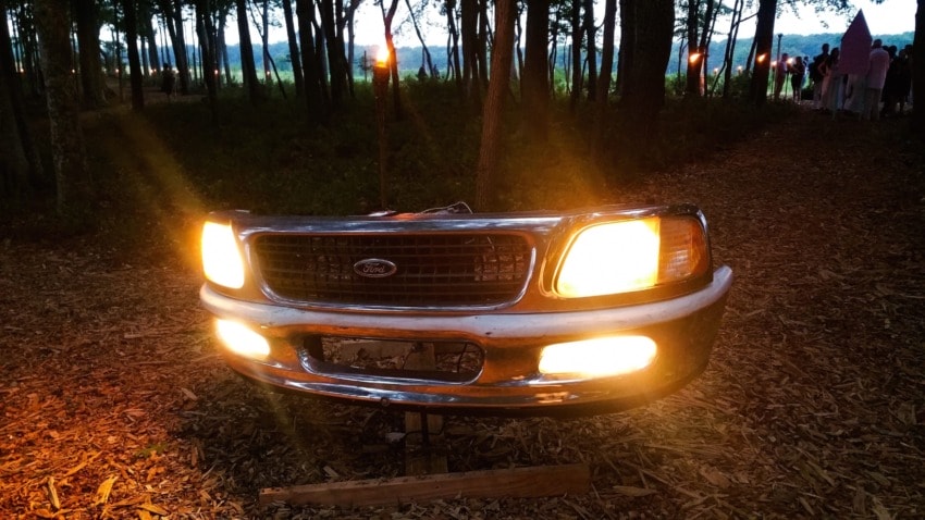 Illuminated car front with headlights in a wooded area at dusk, surrounded by trees and dim sunlight.