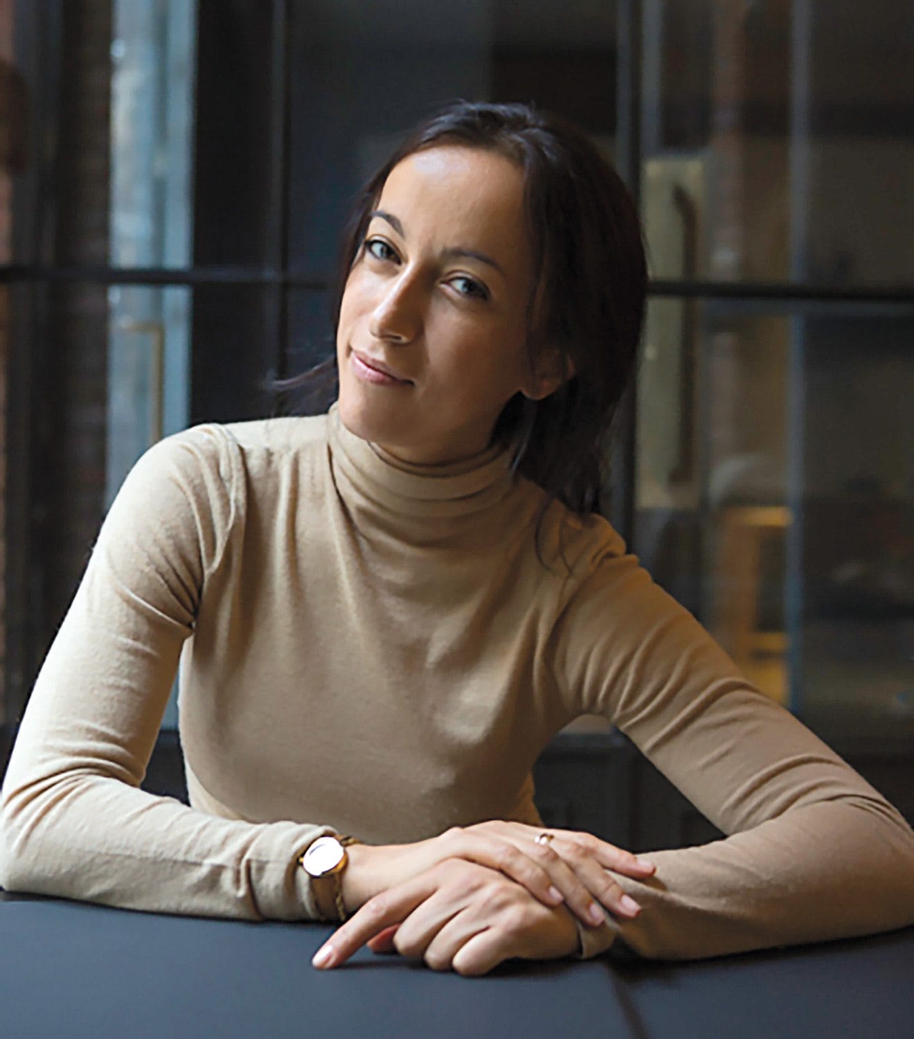 Person sitting at a table wearing a beige turtleneck and watch, with a relaxed expression and hands folded.