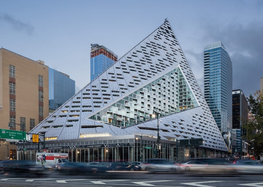 Modern triangular building with unique windows and busy street in foreground, surrounded by skyscrapers at dusk.